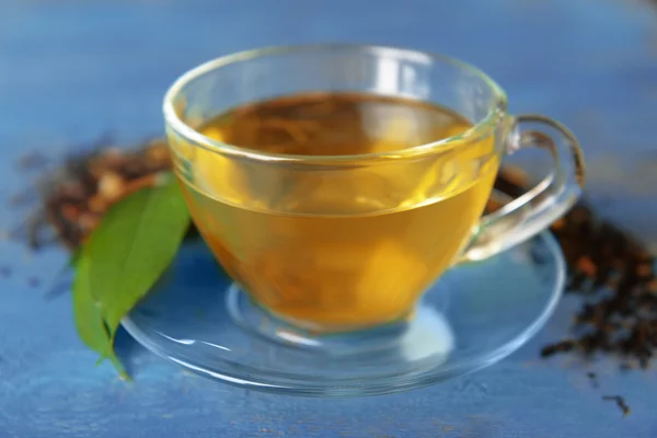 Glass cup of tea with green leaves and scattered tea around on blue wooden background — Stock Photo, Image