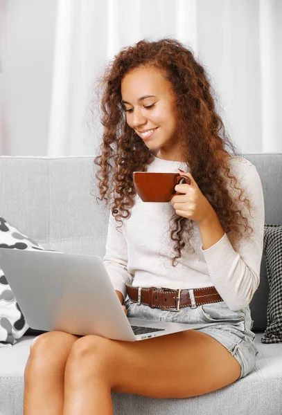 年轻漂亮的女人在沙发上使用笔记本电脑，在房间里喝咖啡 — 图库照片