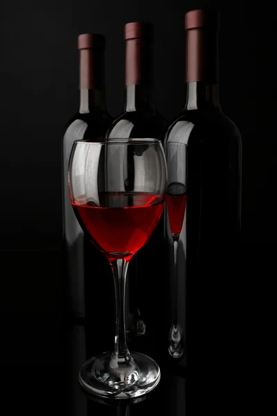 Red wine glass against bottles in a row on black background, close up — Stock Photo, Image