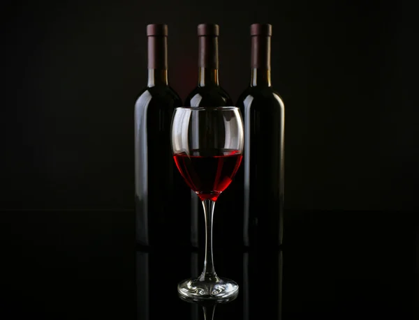 Wine glass against bottles in a row on dark red background, close up — Stock Photo, Image