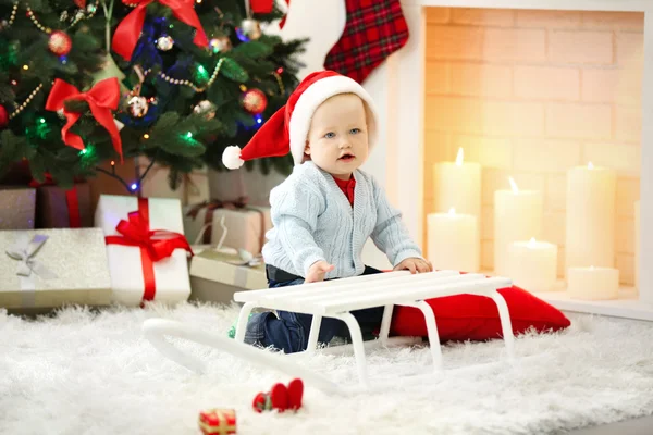 Bebé divertido sentado cerca de trineo y árbol de Navidad y chimenea en el fondo —  Fotos de Stock