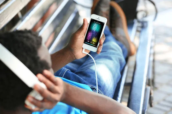 Hombre afroamericano escuchando música con auriculares en el banco en el parque —  Fotos de Stock