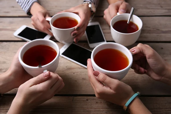 Empat tangan dengan ponsel pintar memegang cangkir dengan teh, di atas meja kayu latar — Stok Foto