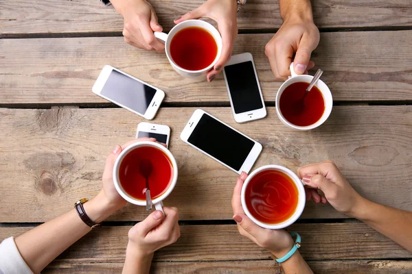 Four hands with smart phones holding  cups with tea, on wooden table background — Stock Photo, Image
