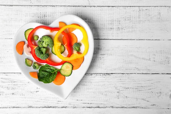 Useful cut vegetables on a plate in the form of heart on wooden table top view — Stock Photo, Image