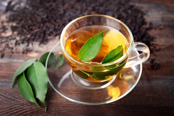 Скляна чашка чаю з зеленим листям на дерев'яному фоні, прикрашена розсіяним чаєм — стокове фото