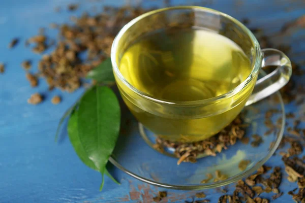 Tazza di tè di vetro con foglie verdi e tè sparso intorno su sfondo di legno blu — Foto Stock