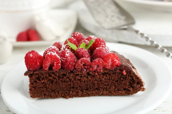 Stück Kuchen mit Schokoladenglasur und Himbeeren auf farbigem Holzhintergrund — Stockfoto