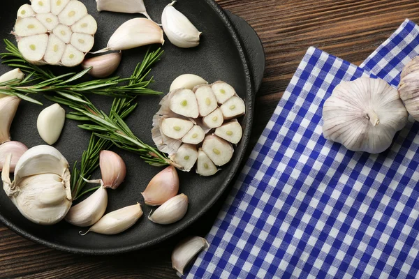 Aglio fresco con rosmarino in padella nera vicino tovagliolo di cotone a quadretti blu su sfondo di legno, primo piano — Foto Stock