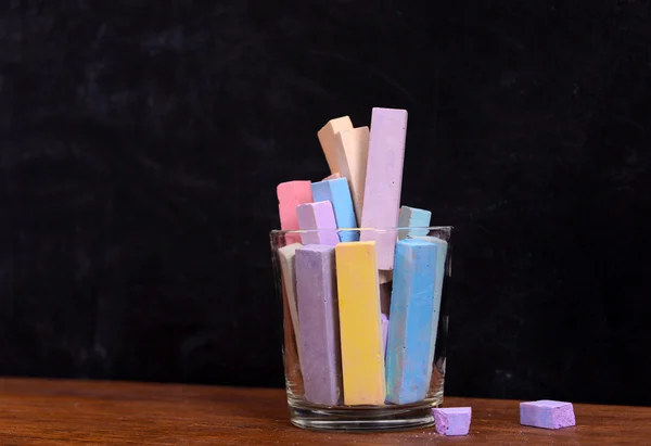 Glass of coloured chalk on desk in front of blackboard — Stock Photo, Image