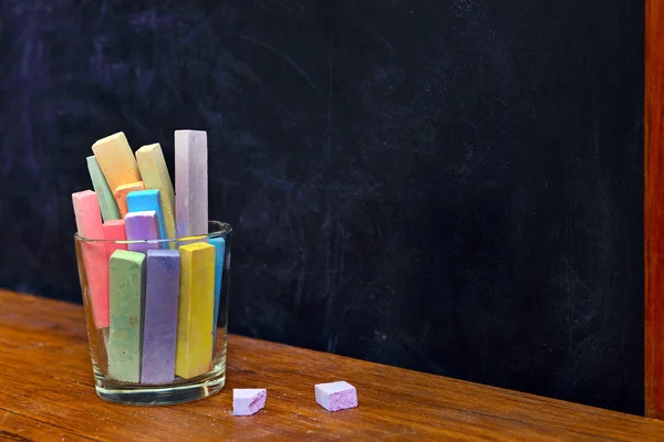 Glass of coloured chalk on desk in front of blackboard — Stock Photo, Image