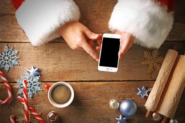 Christmas concept. Santa takes smart phone in hands over wooden table, close up — Stock Photo, Image