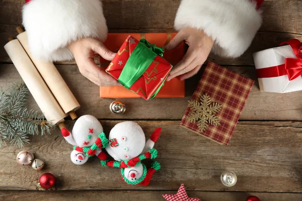 Bella confezione regalo nelle mani di Babbo Natale e decorazioni natalizie su tavolo di legno, primo piano — Foto Stock