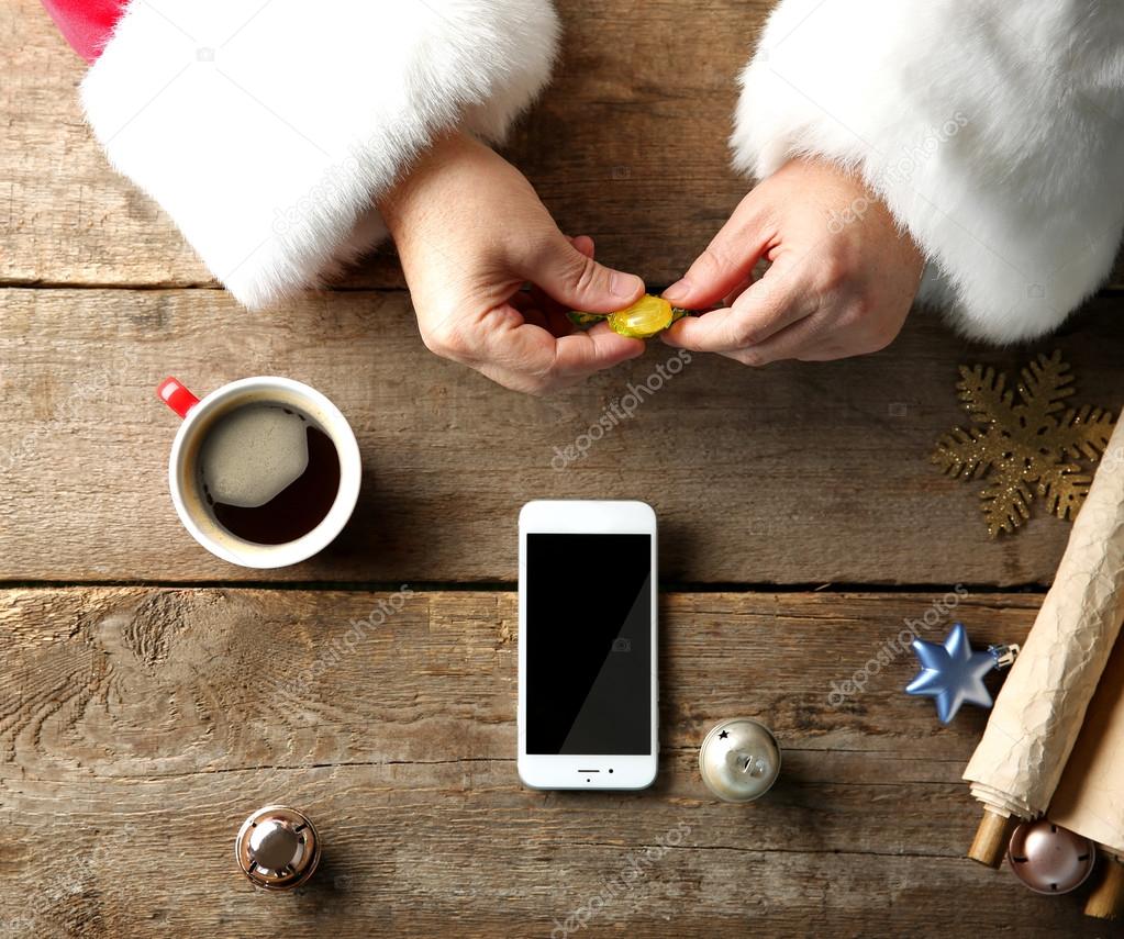 Christmas concept. Santa takes candy in hands. Smart phone, cup of coffee, wish list and decorations on wooden table, close up