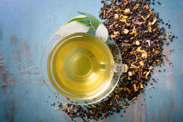 Copa de té de vidrio con hojas verdes y té disperso sobre fondo de madera azul — Foto de Stock