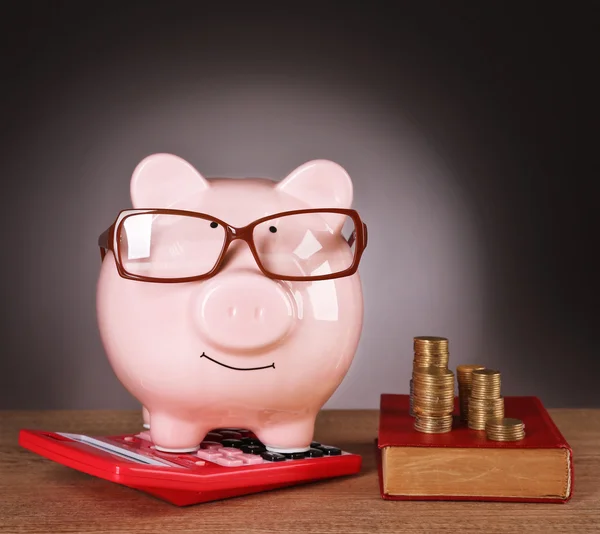 Sparschwein in Brille mit Taschenrechner, Buch und Münzen auf grauem Hintergrund — Stockfoto