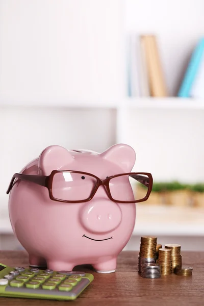 Банк свиней в очках с калькулятором и монетами на домашнем или офисном фоне — стоковое фото