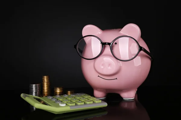 Piggy bank em óculos com calculadora e moedas em fundo escuro — Fotografia de Stock