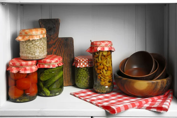 Jars with pickled vegetables and beans, spices, book of recipes and kitchen utensils on shelf — Stock Photo, Image