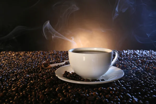 Xícara de café quente entre grãos de café no fundo escuro — Fotografia de Stock