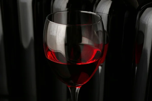 Copo de vinho tinto contra garrafas em uma fileira no fundo preto, close-up — Fotografia de Stock