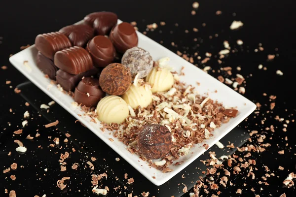 Assorted chocolate candies on white plate with flakes over black background — Stock Photo, Image