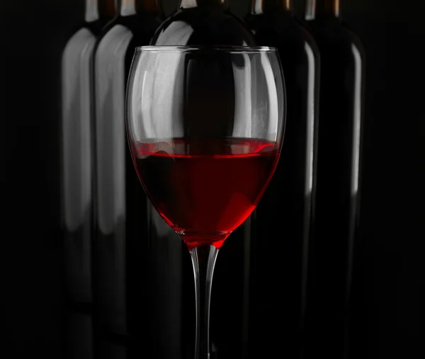 Red wine glass against bottles in a row on black background, close up — Stock Photo, Image