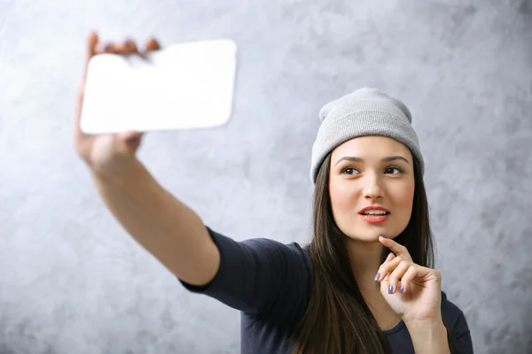 灰色の壁の背景にスマート フォンと彼女の自己の写真を取る帽子の少女 — ストック写真