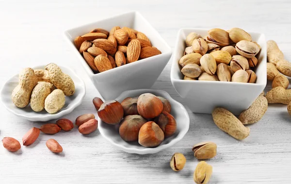 Pistachos, almendras, avellanas, cacahuetes y nueces en los cuencos de cerámica, sobre fondos de madera blanca —  Fotos de Stock