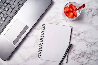 Dizüstü bilgisayar, boş defter ve masada taze dilimlenmiş çilekli bir bardak sağlıklı yoğurt, kopyalama alanı. Freelancer çalışma alanı, merkez ofis. Üst manzara, düz uzanma