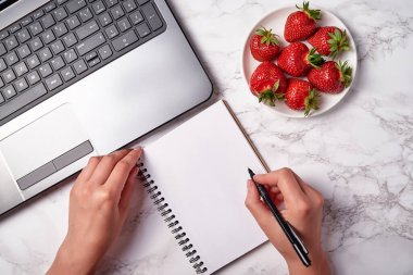 Boş defterle yazan kadın elleri, beyaz masaya dizüstü bilgisayar ve çilekler, kopyalama alanı. Düz yatıyordu