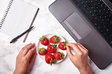 Kadın eli, dizüstü bilgisayar, masadaki boş defter ve çilekler, kopyalama alanı. Freelancer çalışma alanı, merkez ofis. Üst manzara, düz uzanma
