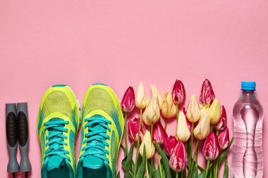 Bir çift spor ayakkabı, ip atlama, bir şişe su ve pembe arka planda laleler. Yeni ayakkabılar, fotokopi alanı. Ayak giysisi çekimleri. Üst manzara, düz uzanma