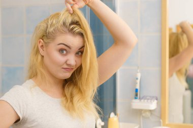 Yağlı saçlarıyla sorunu olan sarışın bir kadın banyoda kendine bakıyor. Kadın kafa derisini gösteriyor, kaşınıyor, kepek problemi var..