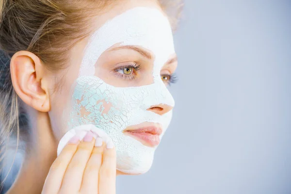 Young woman with white dried mud mask on face being removing cosmetic with cotton swab. Teen girl taking care of oily skin. Beauty treatment. Skincare.