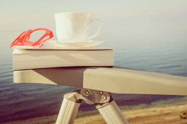 Tatillerde kitap okumak. Kahve fincanı ve kitap açık havada mavi deniz suyuna karşı sandalyede..