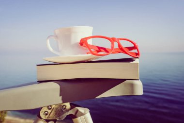 Tatillerde kitap okumak. Kahve fincanı ve kitap açık havada mavi deniz suyuna karşı sandalyede..
