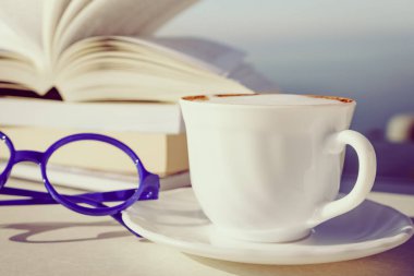 Tatillerde kitap okumak. Kahve fincanı ve kitaplar açık havada mavi deniz suyuna karşı. Kitap sayfaları rüzgarda çırpınıyor.