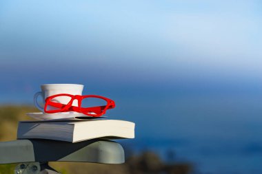 Ruhsal iyileşme, pozitif terapi. Sahile karşı kahve fincanı, kitap ve kırmızı bardaklar. Tatilde okumak. Tatillerde rahatla. Zihinsel sağlık sorunları.