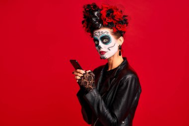 Girl with sugar skull makeup with a wreath of flowers on her head and skull, wearth lace gloves and leather jacket, shocked with news from her phone isolated on red background. clipart
