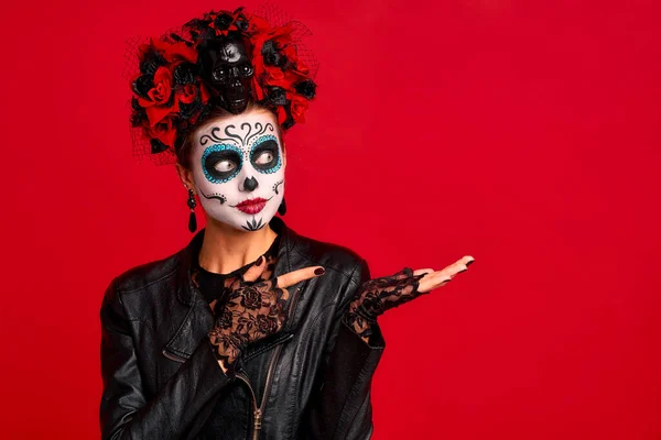Young woman with sugar skull makeup with a wreath of flowers on her head and skull and black gloves points to a free space. isolated on red background. concept of Halloween or Calavera Catrina.