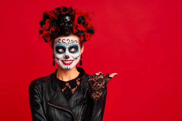 Young woman with sugar skull makeup with a wreath of flowers on her head and skull and black gloves points to a free space. isolated on red background. concept of Halloween or Calavera Catrina.