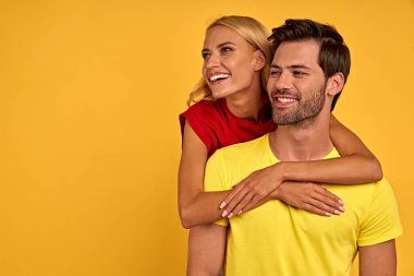 Sarı duvar arkası stüdyo resminde izole edilmiş renkli tişörtler giyen gülümseyen genç çift. İnsanlar yaşam tarzı kavramını hisseder. Kopyalama alanı oluşturun. Sarılmak, yana bakmak.