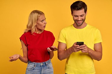 Gülümseyen meraklı genç çift arkadaş tişörtlü kız sarı arka plan stüdyosunda yalnız poz veriyor. İnsanların yaşam tarzı konsepti. Kıskanç bir kız erkek arkadaşının telefonuna bakar..