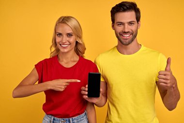 Gülümseyen genç arkadaşlar, sarı arka planda izole edilmiş renkli tişörtlü kız arkadaşlar. İnsanların yaşam tarzı konsepti. Boş ekran ile cep telefonu üzerinde işaret parmağı gösteriliyor, başparmak gösteriliyor