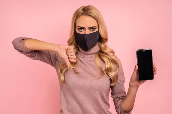 Young girl in pink blouse, face mask isolated on pink background. Epidemic pandemic coronavirus 2019-ncov sars covid-19 flu virus concept Hold mobile phone with blank empty screen showing thumb down.