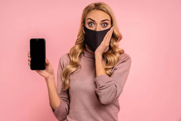 Young woman in pink blouse, face mask to safe from coronavirus virus covid-19 during pandemic quarantine hold mobile cell phone with blank empty screen put hand on head isolated on pink background