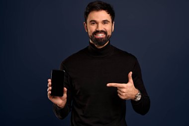 Moda yüksek yakalı süveter giyen yakışıklı adam akıllı telefon sunuyor ve fotokopi alanı olan boş siyah ekranı işaret ediyor. Pasifik mavisi arka planda izole edilmiş..