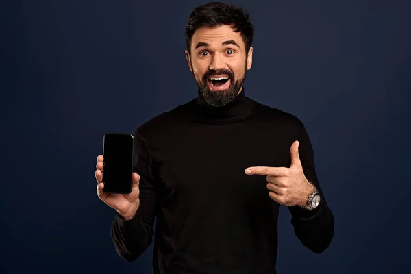 Handsome man in trendy high neck sweater presenting smart phone and pointing with finger at blank black screen with copy space, isolated on pacific Blue background.
