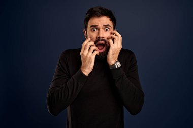 Stüdyoda korkmuş bir adamın paniğe kapılıp ellerini açık tutması, korkunç bir şey görmesi, heyecanlı hissetmesi, siyah kazak giymesi, mavi arka planda izole edilmesi, korkunç haberler duyması..